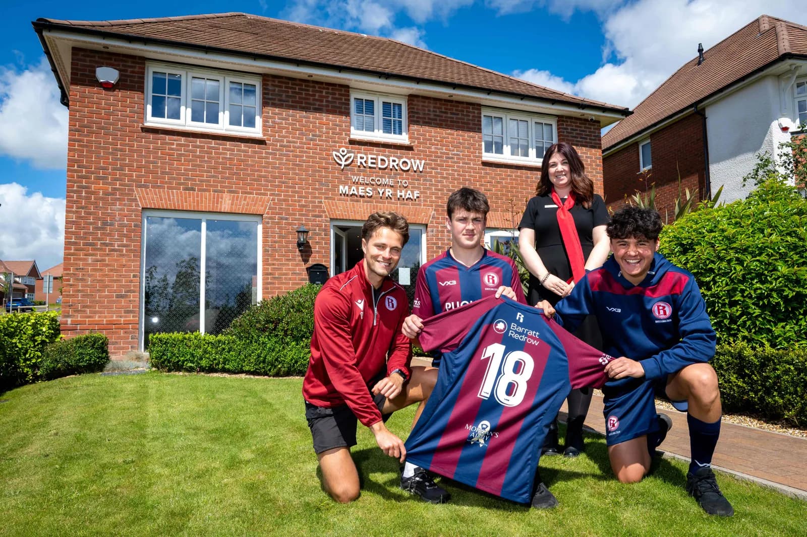 Two young men and an adult man kneeling on grass in front of a red brick building, holding up a red and blue striped soccer jersey with the number 18, while a woman in a black top and red scarf stands behind them.