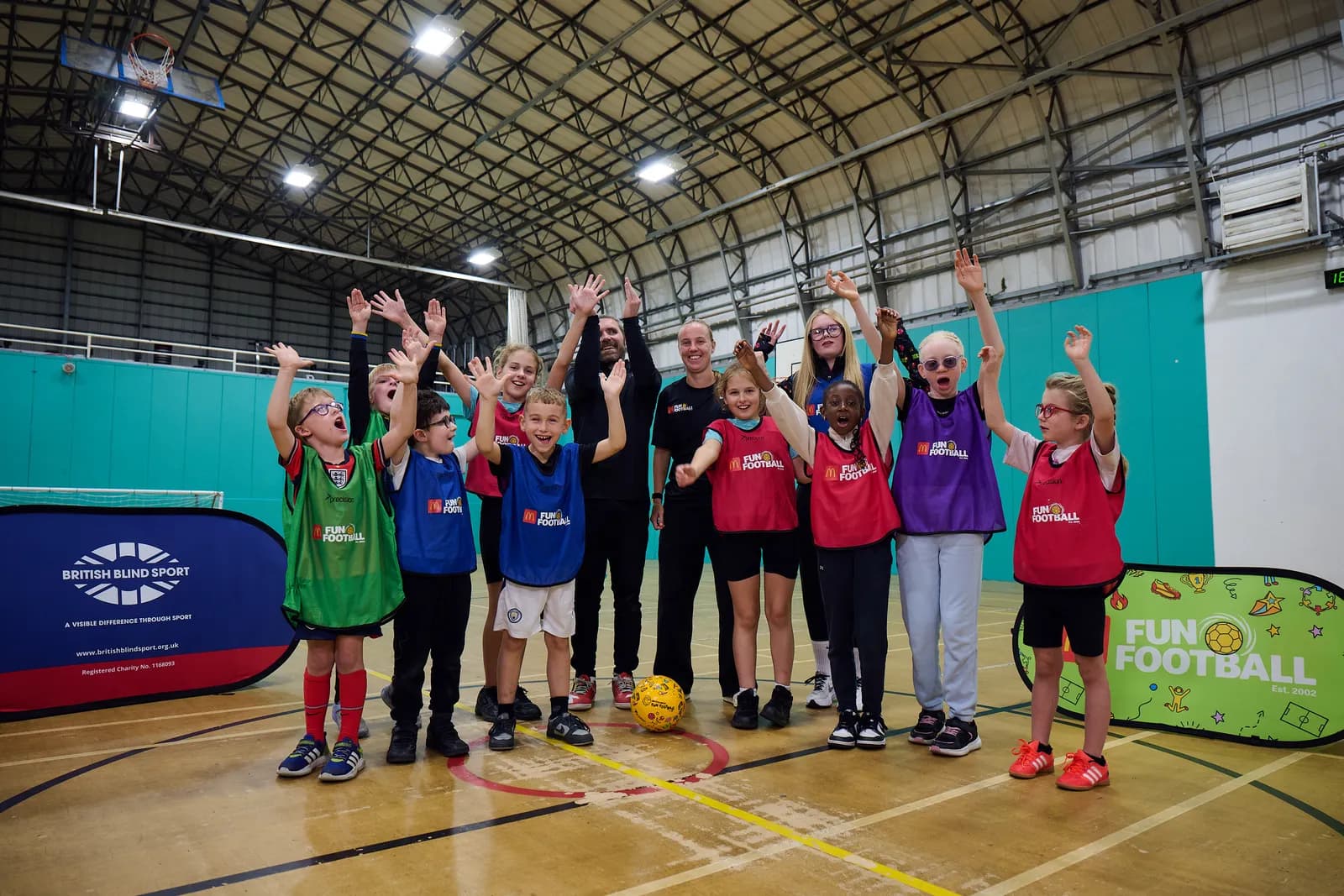 A group of children and adults, all with their hands raised in the air, stand on a basketball court next to a yellow football, with banners for 'British Blind Sport' and 'Fun Football' in the foreground.