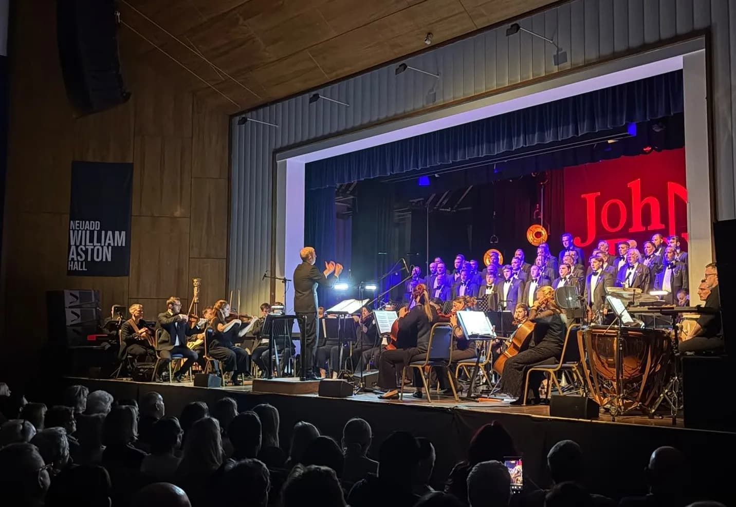 A conductor leads an orchestra and a choir on a stage in front of an audience, with "John" displayed in red lights on the back wall.