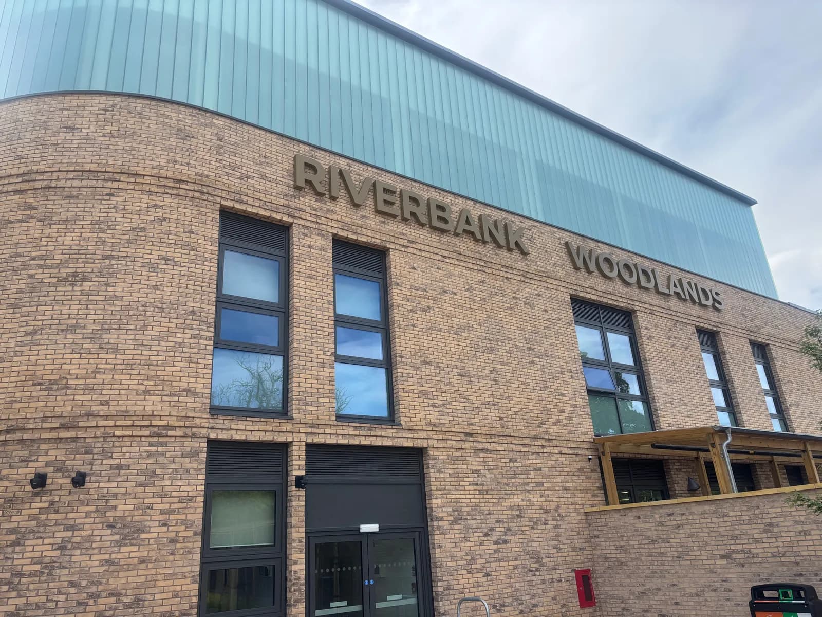 The Riverbank Woodlands building features a brick facade, numerous dark-framed windows reflecting the sky, and a light blue upper level, with the building name in large letters.